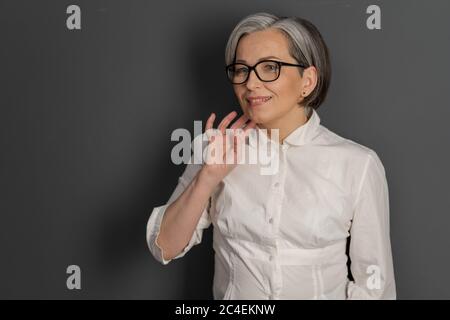 Piangendo bella donna sorride toccando il mento con mano coquettishly. Signora matura intelligente in occhiali e camicia bianca su sfondo grigio. Spazio di testo in Foto Stock