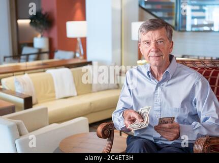 Uomo caucasico senior che tiene alcune fatture del dollaro degli Stati Uniti come se le consegnasse allo spettatore. Composto in un'esclusiva lounge dell'hotel o della nave da crociera Foto Stock