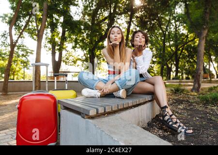 Foto di due attraenti donne turistica soffiando aria kiss mentre è seduto sul banco di lavoro con i bagagli nel parco verde Foto Stock