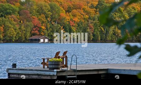 Pensione Living - due sedie Muskoka seduta su una barca dock foglia autunno colore e una boathouse in ba Foto Stock