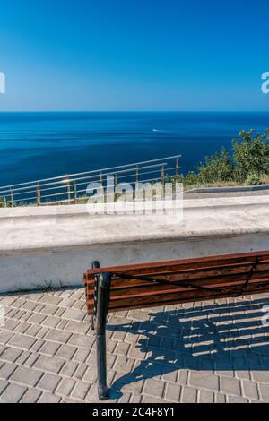 Panca vuota sulla riva del mare in un parco. Vista mare dalla montagna. Concetto di viaggio, relax o solitudine. Panca di legno su spazio aperto che si affaccia su una bella Foto Stock