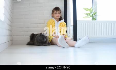 Una ragazza sconvolgita in un accappatoio giallo si trova nell'angolo della camera, un gatto domestico grigio scuro si trova nelle vicinanze Foto Stock