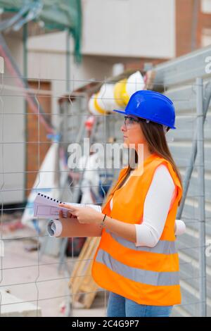 Vista laterale di uno splendido studente di architettura giovane che indossa un equipaggiamento di sicurezza che tiene i progetti e controlla un cantiere. Lavoro e apprendistato c Foto Stock