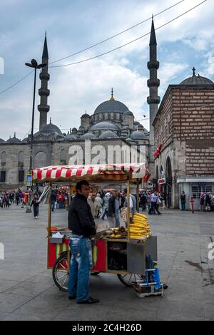 Un uomo si trova accanto al suo carrello mobile di pannocchie nel distretto di Eminonu di Istanbul, in Turchia. Sullo sfondo si trova il 400 anni Yeni Camii. Foto Stock