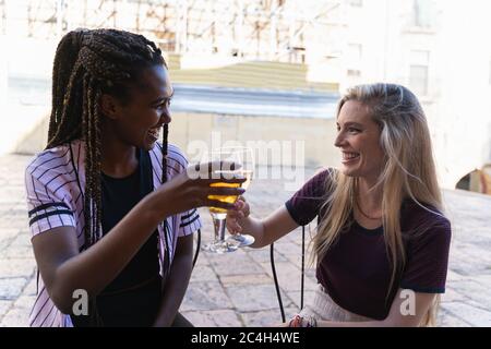 Due giovani fidanzate multietniche che si gustano un drink insieme in un ristorante ridendo e scherzando mentre brindano per celebrare una riunione Foto Stock