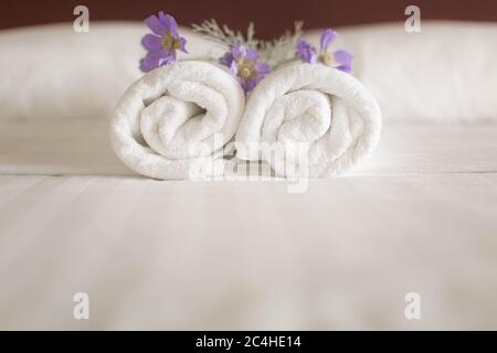 Primo piano di un set di rotoli di asciugamani bianchi su un letto d'hotel con fiori viola su di essi Foto Stock