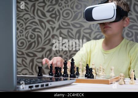 Un ragazzo in una cuffia per realtà virtuale gioca a scacchi con un laptop. Imparare a giocare a scacchi usando la tecnologia informatica Foto Stock