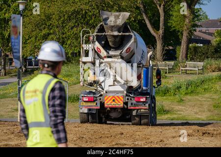 Il lavoratore di costruzione che indossa elmetto e gilet ad alta visibilità orologi quando il camion di calcestruzzo arriva sul cantiere Foto Stock