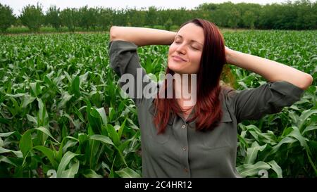 Una donna con i capelli rossi in una camicia verde tiene le mani dietro la testa in un campo. Contadino che sogna in un campo di mais. Foto Stock