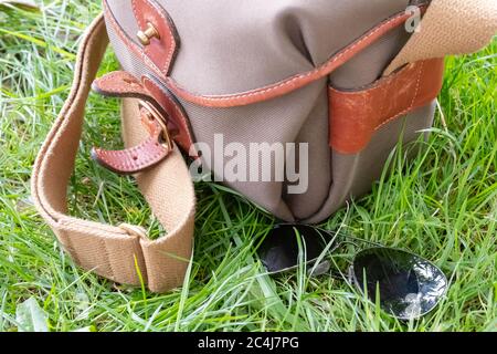 Un paio di occhiali da sole stile aviatore visto adagiato su un'erba lussureggiante accanto a una borsa per macchina fotografica in stile tela. Foto Stock