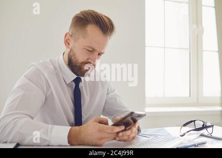 Un uomo d'affari serio e occupato legge il messaggio sul telefono ad un tavolo con un calcolatore nell'ufficio. Foto Stock