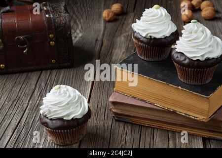 I cupcake in cartoni di carta paty, sormontati con panna montata, sono giacenti su libri antichi e tavole di legno stagionato. Un petto e qualche guscio di noce... Foto Stock