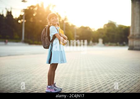 Una scolaretta con uno zaino e libri si trova su una strada della città. Ritorno a scuola. Foto Stock