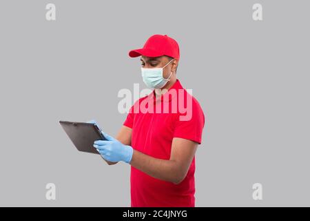 Uomo di consegna che usa la compressa che indossa la maschera medica e guanti. Ragazzo di consegna indiano con la compressa in mani. Consegna a domicilio, consegna tecnica Foto Stock