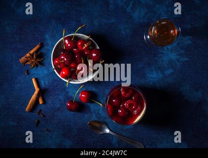 Panna cotta dessert tradizionale italiano con ciliegie in camicia al rum e sciroppo di miele. Tavolo di fondo blu scuro, bicchiere di rum, ciotola di ciliegie fresche Foto Stock