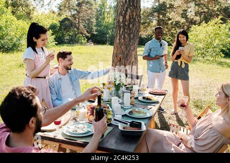 Tre giovani coppie felici in casualwear godendo di una giornata di sole, mentre uno di loro scorrendo nel loro smartphone da pino albero su erba verde Foto Stock