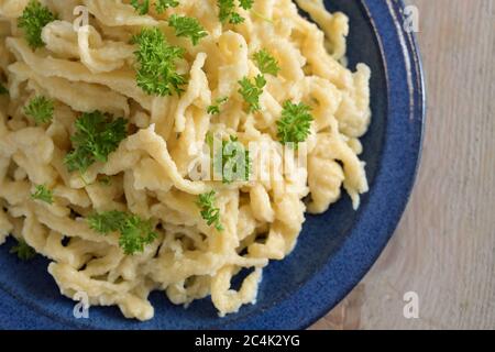 Spaghetti tedeschi fatti in casa con formaggio, chiamati kaes spaetzle, serviti con contorno di prezzemolo su un piatto blu e un rustico tavolo in legno, vista ad angolo alto Foto Stock