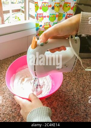 Primo piano foto di una donna che mescola crema per cuocere una torta usando un miscelatore elettrico con una ciotola rosa. Concetto di cucina a casa. Le attività interne, più realistiche Foto Stock