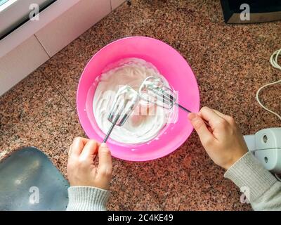 Primo piano foto di una donna che mescola crema per cuocere una torta usando un miscelatore elettrico con una ciotola rosa. Concetto di cucina a casa. Le attività interne, più realistiche Foto Stock