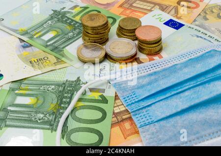 Pile di monete e banconote in euro con maschera medica. Pandemia di coronavirus in Europa. Impatto finanziario della COVID-19 nell'UE. Foto Stock