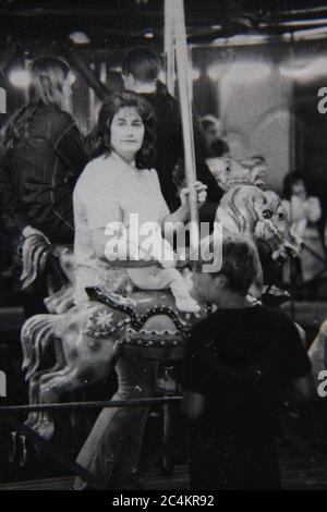 Bella fotografia di stile di vita in bianco e nero anni '70 di una madre che prende un giro sulla giostra al carnevale di tarda notte. Foto Stock