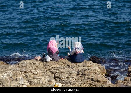 Scatto ad angolo alto di due femmine che siedono sulle rocce vicino il mare mentre indossa hijab Foto Stock