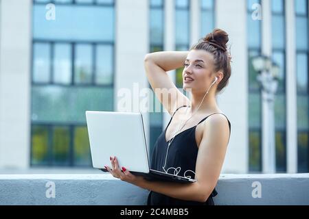 bella ragazza cammina per la città, lavorando su un computer portatile, tenendo un computer portatile e ascoltando musica. Foto Stock