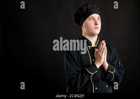 Ritratto di giovane maschio vestito in un vestito nero dello chef sta pregando in posa su uno sfondo nero isolato con area di pubblicità dello spazio di copia Foto Stock
