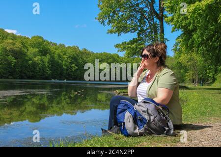 Donna escursionista seduto sul Lake Edge Bank con zaino e. Sigaretta per fumatori con occhiali da sole Foto Stock
