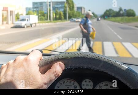 Vista dalla vettura, la mano dell'uomo al volante della vettura, che si trova di fronte alla zona pedonale e pedoni che attraversano la strada Foto Stock