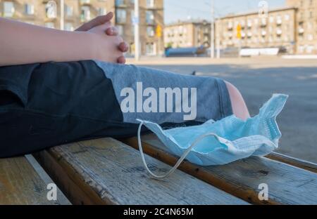 La maschera medica antibatterica abbandonata e usata si trova sull'asfalto ai piedi di un uomo che è seduto su una panchina in un parco cittadino Foto Stock