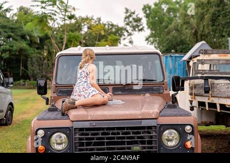 Townsville, Queensland, Australia - Giugno 2020: Giovane bionda caucasica con capelli lunghi si siede sul cofano di una vettura d'epoca nel suo cortile Foto Stock