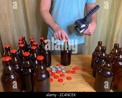 Birra artigianale in casa, l'uomo chiude le bottiglie di birra di vetro marrone con il cappero di plastica su un tavolo di legno con i tappi a corona rossi. Immagine orizzontale di scorta Foto Stock