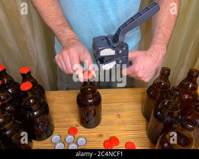 Birra artigianale in casa, l'uomo chiude le bottiglie di birra di vetro marrone con il cappero di plastica su un tavolo di legno con i tappi a corona rossi. Immagine orizzontale di scorta Foto Stock