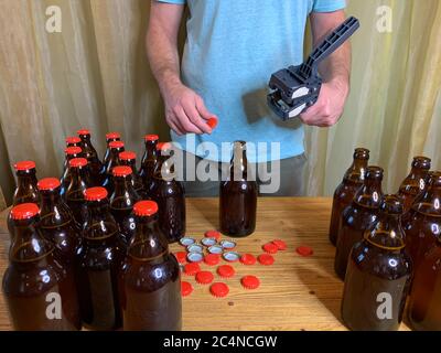 Birra artigianale in casa, l'uomo chiude le bottiglie di birra di vetro marrone con il cappero di plastica su un tavolo di legno con i tappi a corona rossi. Immagine orizzontale di scorta Foto Stock