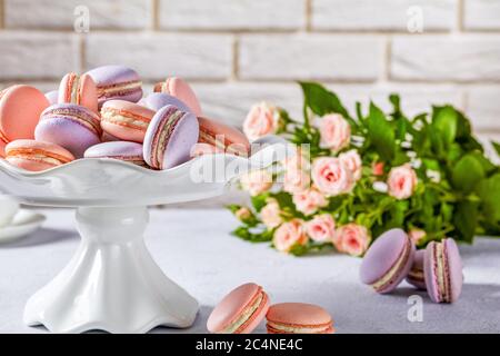 macaron su un supporto per torte in porcellana bianca. bel bouquet di rose e muro di mattoni sullo sfondo, primo piano Foto Stock