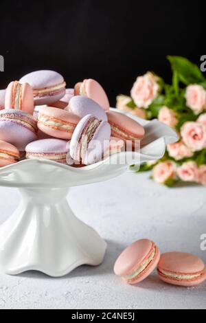 macaron rosa e lavanda su un supporto per torte in porcellana bianca con un bel bouquet di rose su sfondo nero, vista verticale, primo piano Foto Stock