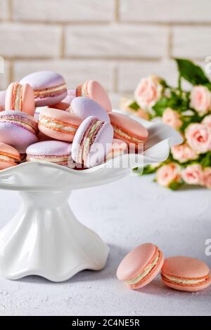 macaron rosa e lavanda su un supporto per torte in porcellana bianca con un bel bouquet di rose sullo sfondo della parete in mattoni, vista verticale, primo piano Foto Stock