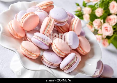 macaron francesi su un supporto per torte in porcellana con un bel bouquet di rose e crema sullo sfondo, vista orizzontale, primo piano, macro Foto Stock