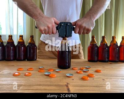 Birra artigianale in casa, l'uomo chiude le bottiglie di birra di vetro marrone con il cappero di plastica su un tavolo di legno con i tappi di corona arancione. Immagine orizzontale Foto Stock