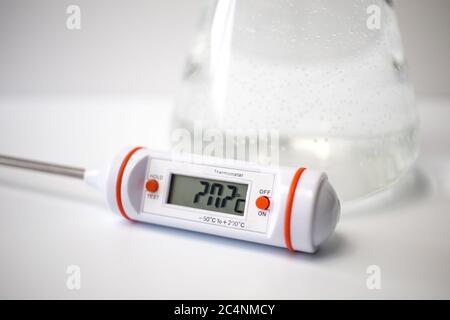Termometro digitale davanti a un liquido in un matraccio di erlenmeyer in vetro su fondo bianco. Esperimento scientifico. Foto Stock