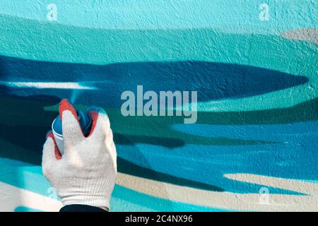 primo piano della mano di un uomo in un guanto tiene un palloncino con vernice e dipinge il muro in blu e bianco Foto Stock