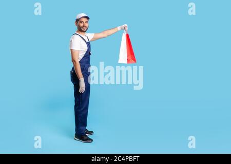 Il corriere professionale felice a lunghezza intera in uniforme tiene le borse della spesa e sorridente alla macchina fotografica, trasportando il pacco con le merci ordinate in linea a. Foto Stock