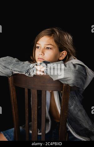 Ragazzo di undici anni con felpa con cappuccio e giacca jean seduta su una sedia di legno su sfondo nero Foto Stock