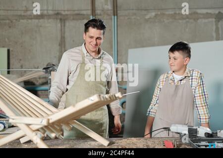 Sorridente padre e figlio di mezza età in grembiuli guardando la sedia di legno in officina di Falegnameria Foto Stock