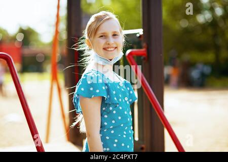 Vita durante la pandemia di covid-19. Ritratto di bambino moderno sorridente in tuta blu con maschera medica sul parco giochi all'aperto della città. Foto Stock