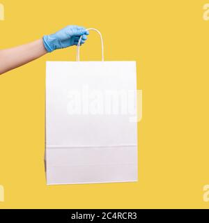 Profilo Vista laterale primo piano della mano umana in guanti chirurgici blu che tengono e mostrano la borsa da shopping bianca. Interno, studio shot, isolato su sfondo giallo Foto Stock