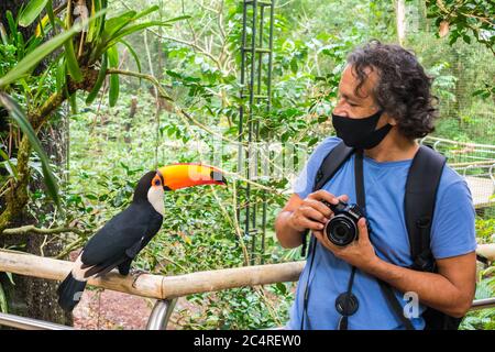 Foz do Iguacu, Brasile - 11 Giugno, 2020: Turistico con una maschera protettiva che interagisce con un curioso toucan al Parco degli Uccelli, popolare destinazione turistica Foto Stock