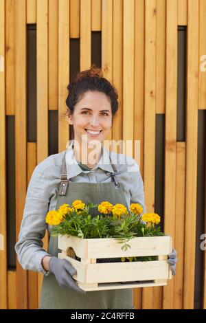 Verticale vita su ritratto di allegra donna giardiniere che tiene i fiori e guardando la macchina fotografica mentre si sta in piedi da sfondo grafico di legno Foto Stock