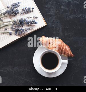 Tazza di caffè con croissant, libro e bouquet di lavanda su tavola nera. Vista dall'alto, disposizione piatta Foto Stock